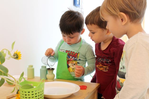 Ein Kind schenkt Wasser in eine kleine Vase, während zwei andere Kinder dabei zusehen